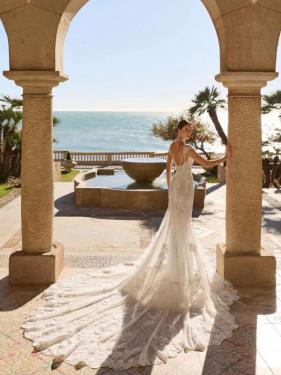 Zu sehen ist ein weißes, figurbetontes Brautkleid aus Spitze mit tiefem Rückenausschnitt und feinen Trägerdetails. Das Kleid hat eine sehr lange, breite Spitzenschleppe.